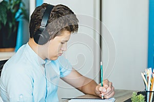 Cute young boy in blue shirt sitting behind desk in his room next to laptop and study. Teenager in earphones makes homework,