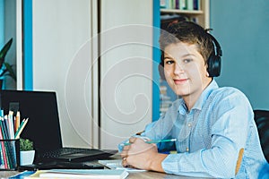 Cute young boy in blue shirt sitting behind desk in his room next to laptop and study. Teenager in earphones makes homework,