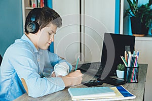 Cute young boy in blue shirt sitting behind desk in his room next to laptop and study. Teenager in earphones makes homework,