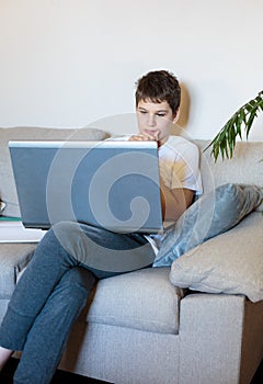 Cute young boy in glasses and white t shirt sitting on the couch in the living room with laptop and study. Homeschooling,