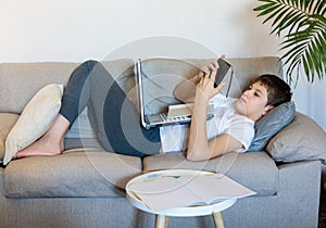 Cute young boy in glasses and white t shirt sitting on the couch in the living room with laptop and study. Homeschooling,