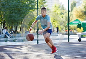 A cute young boy plays basketball on the street playground in summer. Teenager in a green t-shirt with orange basketball ball