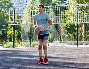 A cute young boy plays basketball on the street playground in summer. Teenager in a green t-shirt with orange basketball ball