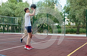 Cute young boy plays basketball on street playground in summer. Teenager in white t-shirt with orange basketball ball outside.