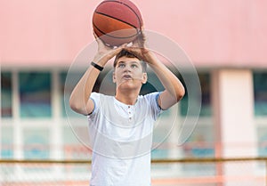 A cute young boy plays basketball on the street playground in summer. Teenager in a white t-shirt with orange basketball ball