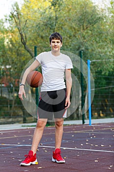 Cute young boy plays basketball on street playground in summer. Teenager in white t-shirt with orange basketball ball outside.