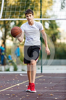 Cute young boy plays basketball on street playground in summer. Teenager in white t-shirt with orange basketball ball outside.