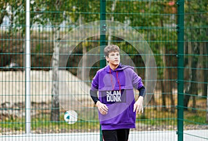 Cute young boy plays basketball on street playground. Teenager in purple hoodie with orange basketball ball outside. Hobby,