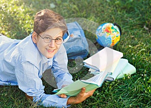 Cute, young boy in round glasses and blue shirt reads book while lying on the grass in the park. Education, back to school