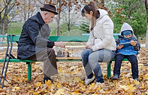 Cute young boy sitting on a park bench holding a tablet computer while his mother and grandfather play chess