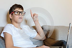 Cute young boy white t shirt sitting on the couch in the living room with his finger up next to laptop and study. Home education,