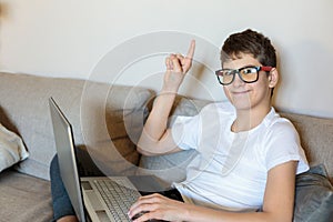 Cute young boy white t shirt sitting on the couch in the living room with his finger up next to laptop and study. Home education,