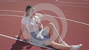 Cute young brunette girl dressed in sneakers, a skirt and a T-shirt sits at outdoor basketball court holding a tennis