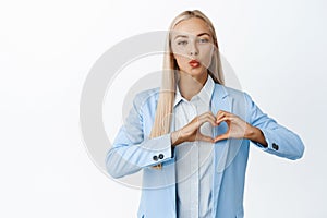 Cute young business woman in suit, showing heart gesture and pucker lips, kissing, standing against white background