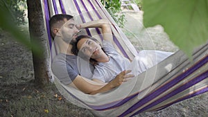 Cute young caucasian man and woman lying in hammock in the garden relaxing together. Loving couple together outdoors