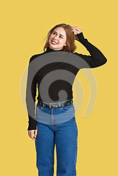 Cute young Caucasian woman with a puzzled and funny expression stands scratching the back of her head. Yellow background