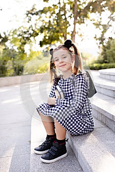 Cute young child girl 5-6 year old wear checkered black and white dress and backpack holding books sitting on stairs outdoors clos