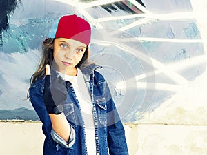 Cute young fair-haired girl teenager in a baseball cap and denim shirt on a stone wall background.