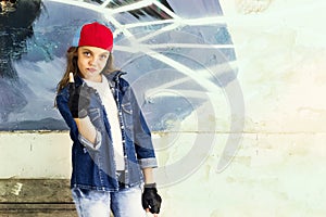 Cute young fair-haired girl teenager in a baseball cap and denim shirt on a stone wall background.