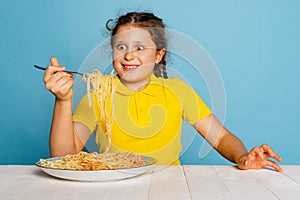 Cute young girl, emotive kid eating delicious Italian pasta isolated on blue studio background. World pasta day