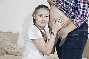 Cute young girl hugging her pregnant mom`s tummy, looking at camera and smiling