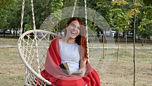 Cute young girl resting on a white swing in the city park. She reads a book and laughs provocatively on a sunny autumn