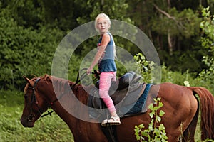 Cute young girl riding a brown horse on a country road at sunset. The child quietly travels on a stallion. The girl confidently