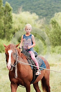 Cute young girl riding a brown horse on a country road at sunset. The child quietly travels on a stallion. The girl confidently