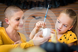 Cute young girl sitting at a table, decorating little white pumpkins with her mother, a cancer patient. DIY Halloween.