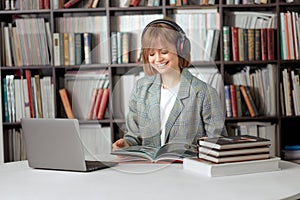 Cute young girl student working on a University project in the library on a laptop, with a book and headphones.