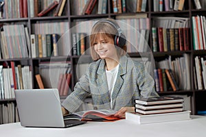 Cute young girl student working on a University project in the library on a laptop, with a book and headphones.