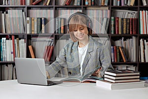 Cute young girl student working on a University project in the library on a laptop, with a book and headphones.