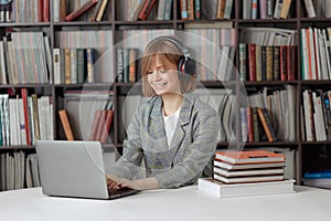 Cute young girl student working on a University project in the library on a laptop, with a book and headphones.