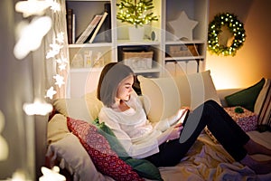 Cute young girl using a tablet pc at home in warm and cozy living room at Christmas. Family having online video call on Xmas eve