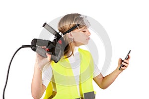 Cute young girl wearing safety vest and safety goggles, holding a rotary hammer and using a smartphone.