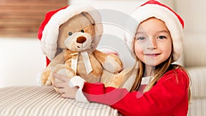 Cute young girl wearing santa hat hugging her christmas present, soft toy teddy bear. Happy kid with xmas present, smiling.