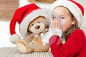 Cute young girl wearing santa hat whispering a secret to her teddy bear christmas present toy. Cheeky kid with teddy bear.