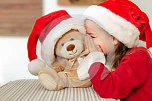 Cute young girl wearing santa hat whispering a secret to her teddy bear christmas present toy. Kid sharing secrets with teddy bear