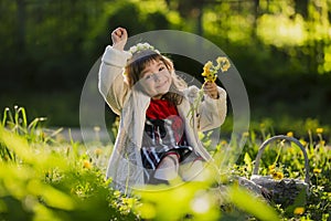 Cute young girl wearing wreath of dandelions and smiling while sitting on grass in park