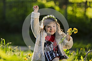 Cute young girl wearing wreath of dandelions and smiling while sitting on grass in park