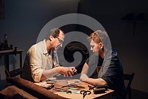 A cute young guy wants to make a gift with his own hands, an experienced shoemaker helps him make shoes out of leather.