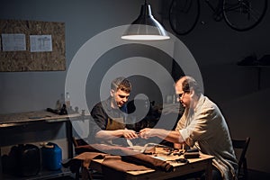 a cute young guy wants to make a gift with his own hands, an experienced shoemaker helps him make shoes out of leather.