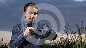 A cute young man checks the messages on a smartwatch at sunset on the field. Beautiful sky on a background.