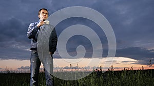 A cute young man drinks a hot drink at sunset on the field. Beautiful sky on a background.