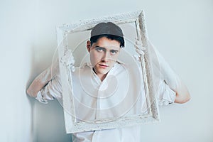 A cute young man wearing a white shirt, holding a blank white frame for a painting. The role of the artist in creating something