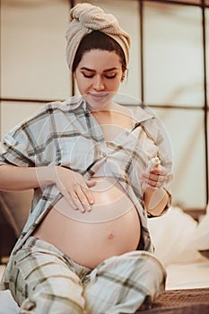 Cute young pregnant woman wearing pajama and wrapped towel on head applying body lotion on her belly in shape of heart and smiling