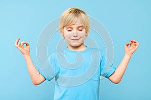 Cute young preschooler boy in yoga position, meditating isolated over pastel blue background. Calm, relax, meditation.