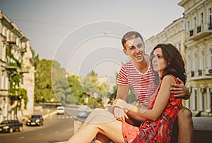 Cute young smiling couple in love hugging, sitting outdoors at green city street, summertime. Girl is wearing glasses of her boyfr