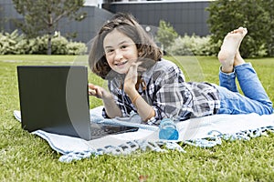 A cute young teenage girl lies on the grass and working on a laptop. Distance learning and education concept. A smiling happy