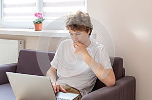 Cute young teenager with laptop on the couch at home. Boy makes homework with a computer. Distance, online education